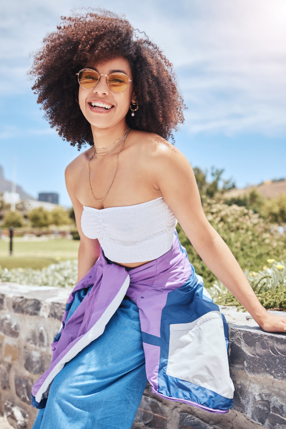A smiling woman with curly hair and sunglasses sitting on a low wall by the sea.